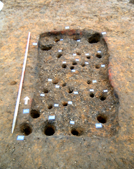 EF62 bustum, pre-excavation and fully-excavated. The stake-holes are presumably the remains of a wooden structure to support the fuel and body.