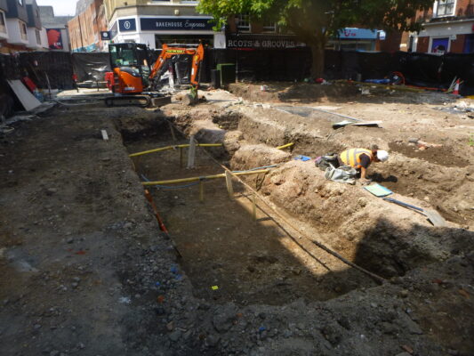 Ongoing archaeological work on St Nicolas Square with an archaeologist recording and a machine in the background.