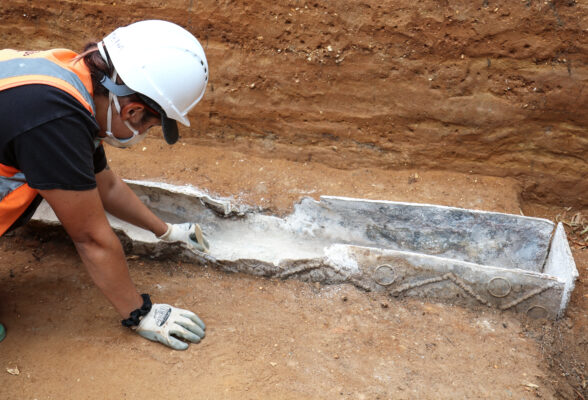 Female archaeologist cleaning out the bas eof a late Roman lead coffin in mask, gloves and hardhat